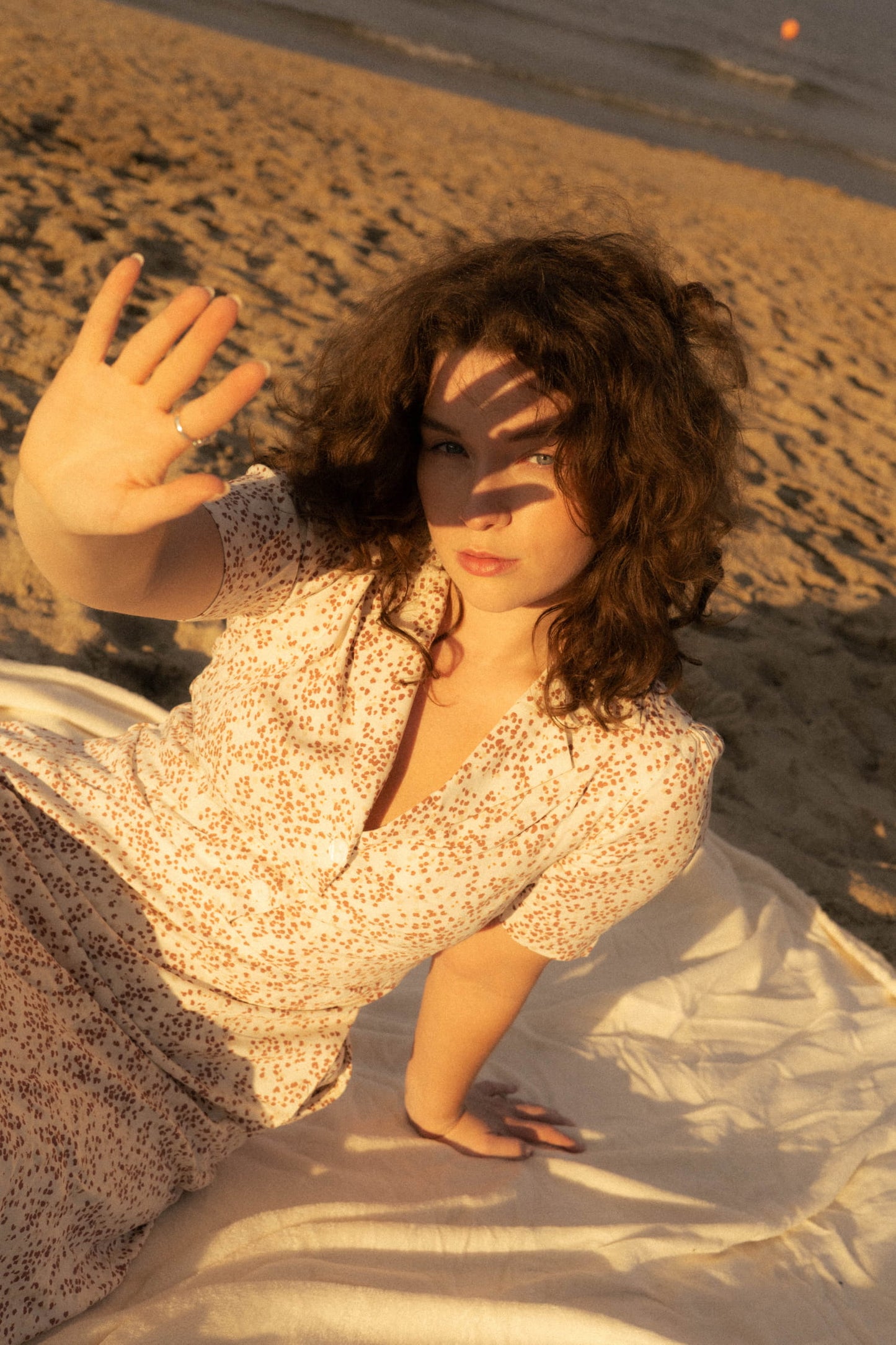 Modelka w sukience Bellucci w print Rosella, siedząca na plaży i osłaniająca się przed zachodzącym słońcem.