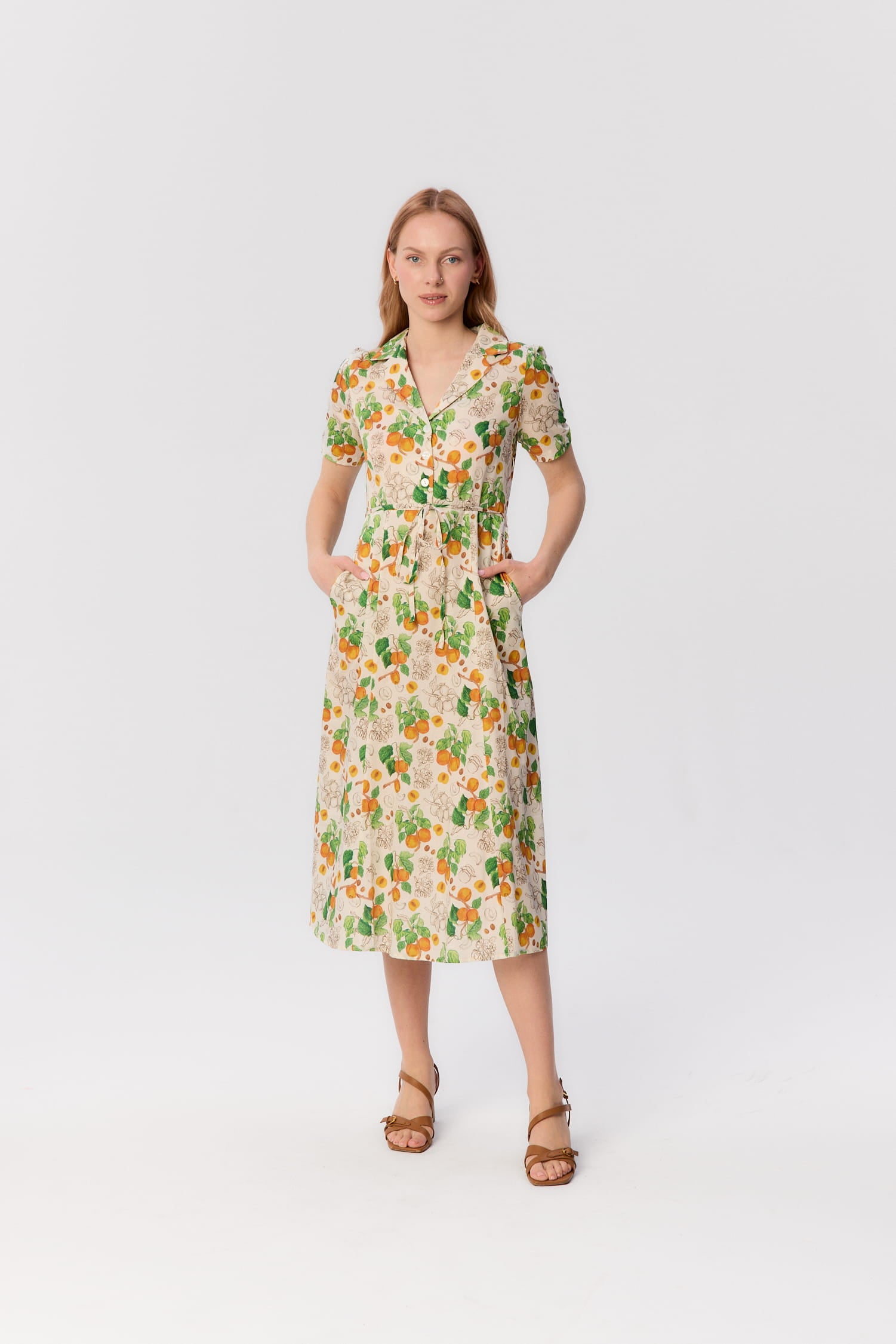 Modelka w sukience Bellucci z satyny bawełnianej w print moreli – krój z kołnierzykiem, krótkim rękawem, guziki i pasek w talii, dłonie w kieszeniach.