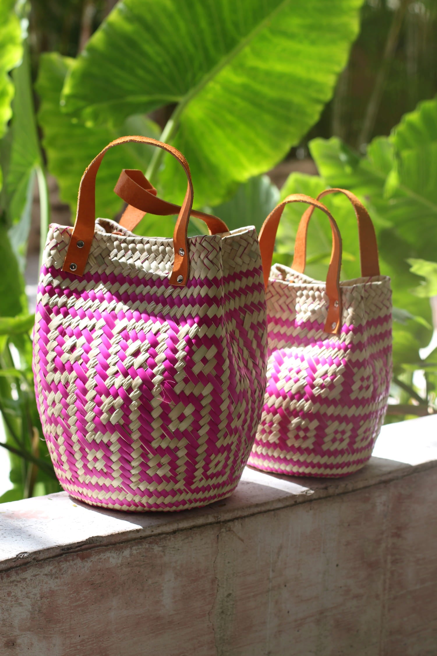 Ręcznie plecione koszyki Vitti w odcieniach różu, z rączkami ze skóry, sfotografowane w promieniach słońca na tle dużych zielonych liści – styl boho, rękodzieło z Meksyku.