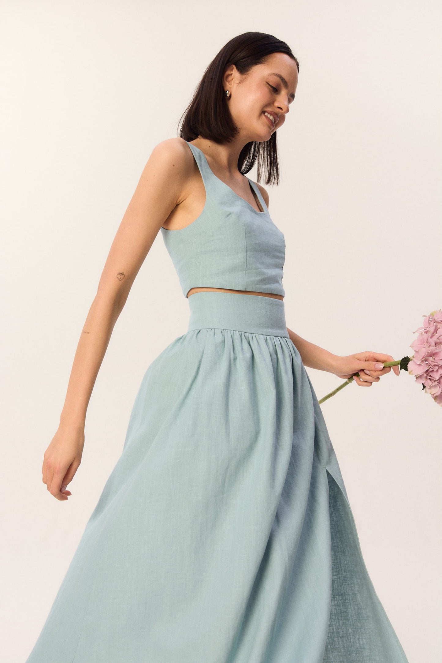 Modelka w turkusowym komplecie MAG NOLLY – top Milo i spódnica Sandrelli z rozcięciem, trzymająca różową hortensję.