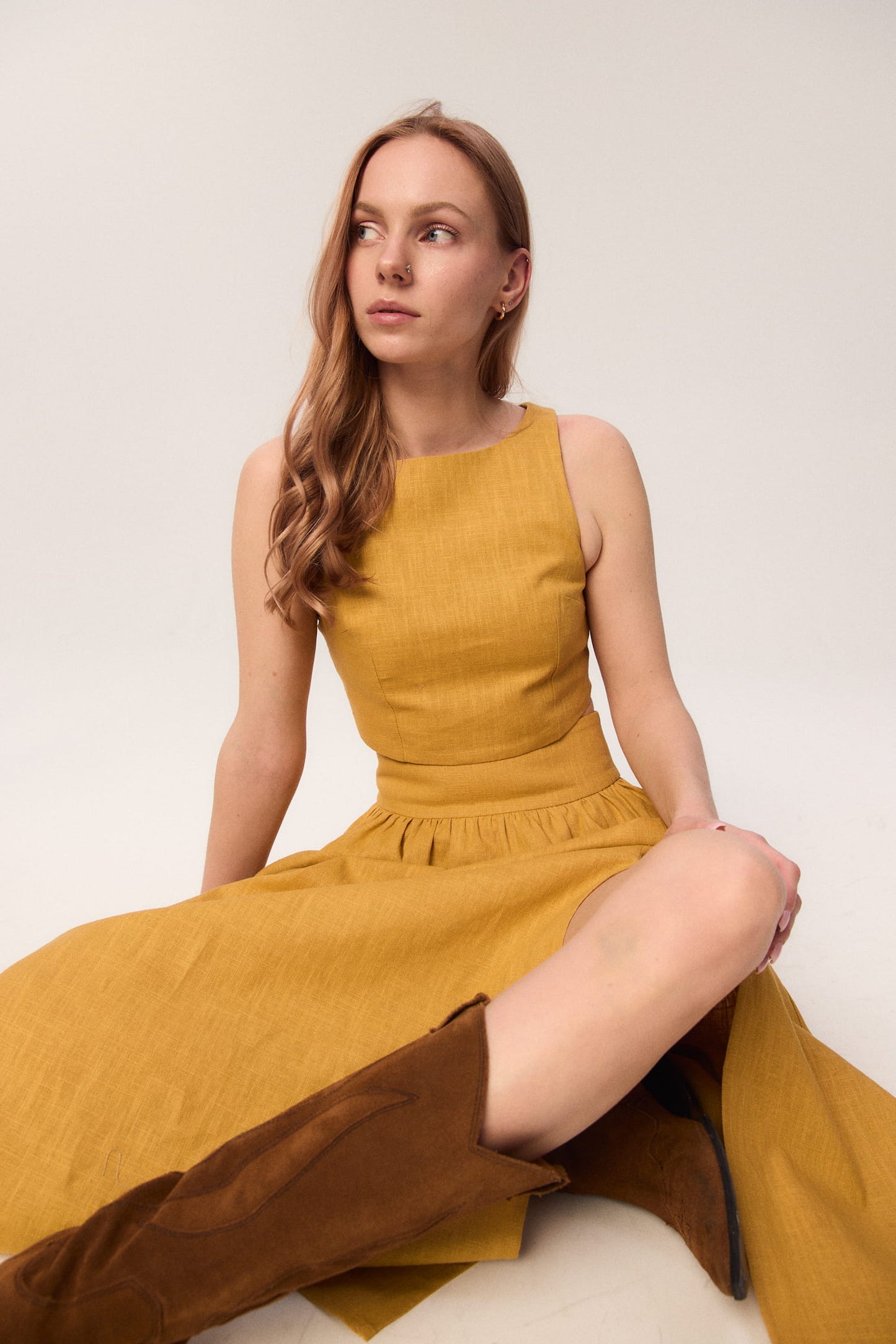 Modelka w zestawie MAG NOLLY – spódnicy Sandrelli i topie Loren w kolorze ochry, w pozycji siedzącej, stylizacja z kowbojkami.