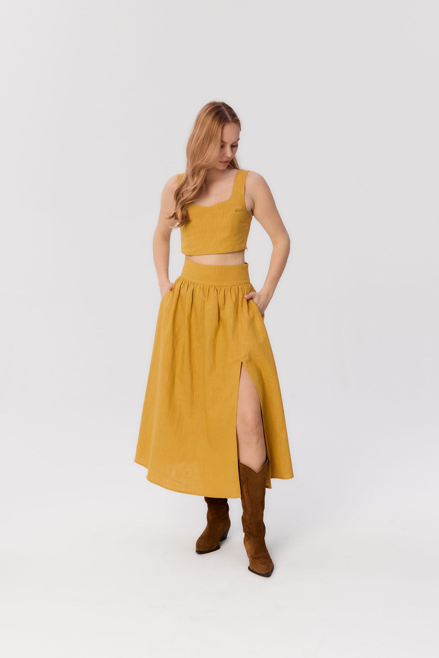 Modelka w zestawie MAG NOLLY – spódnicy Sandrelli i topie Milo w kolorze ochry, stojąca, z rękami w kieszeniach, w kowbojkach.