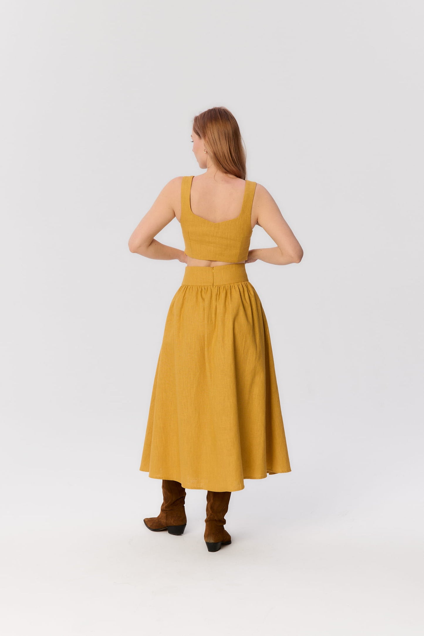 Modelka tyłem w zestawie MAG NOLLY – spódnicy Sandrelli i topie Milo w kolorze ochry, w brązowych kowbojkach.