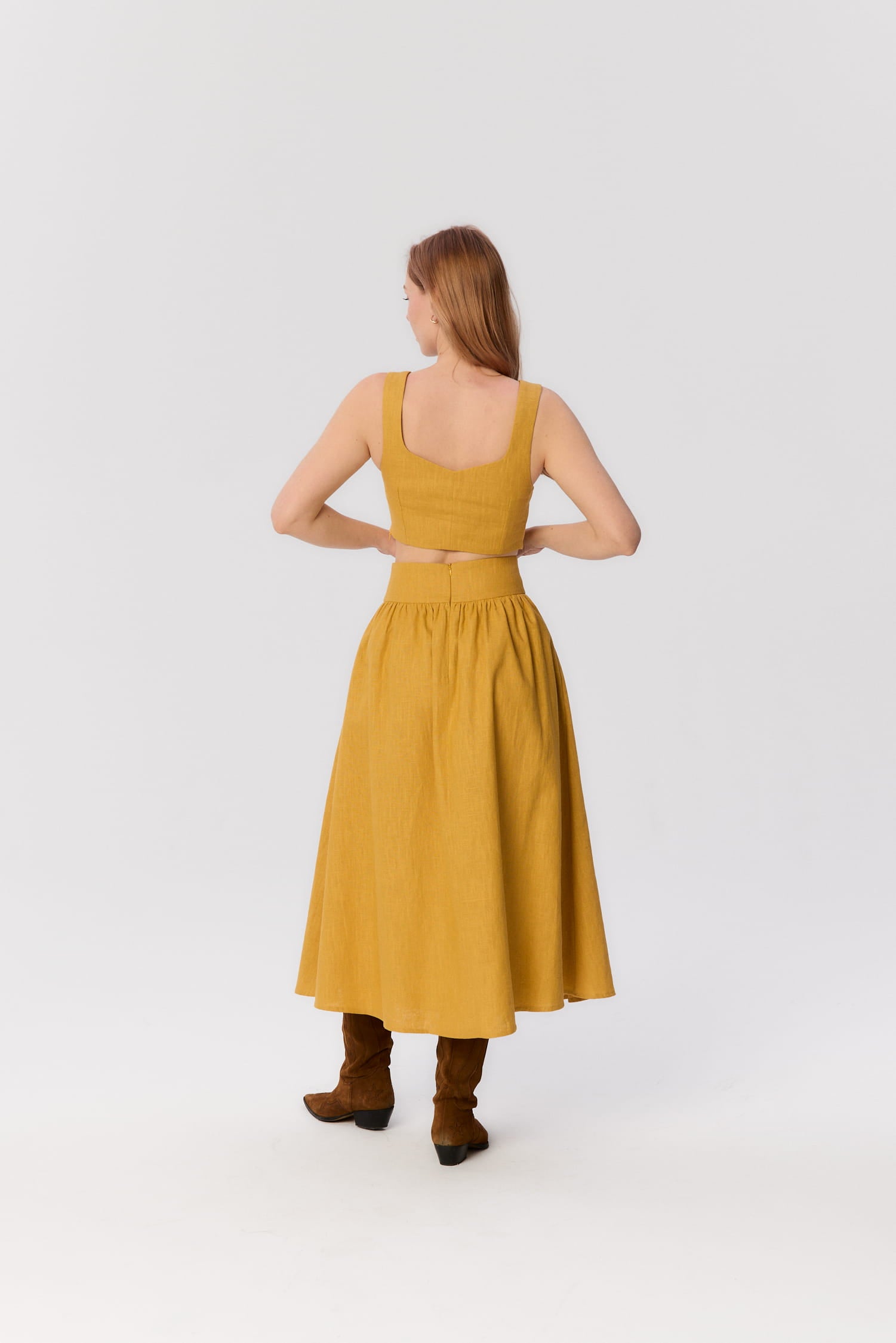 Modelka tyłem w zestawie MAG NOLLY – spódnicy Sandrelli i topie Milo w kolorze ochry, w brązowych kowbojkach.