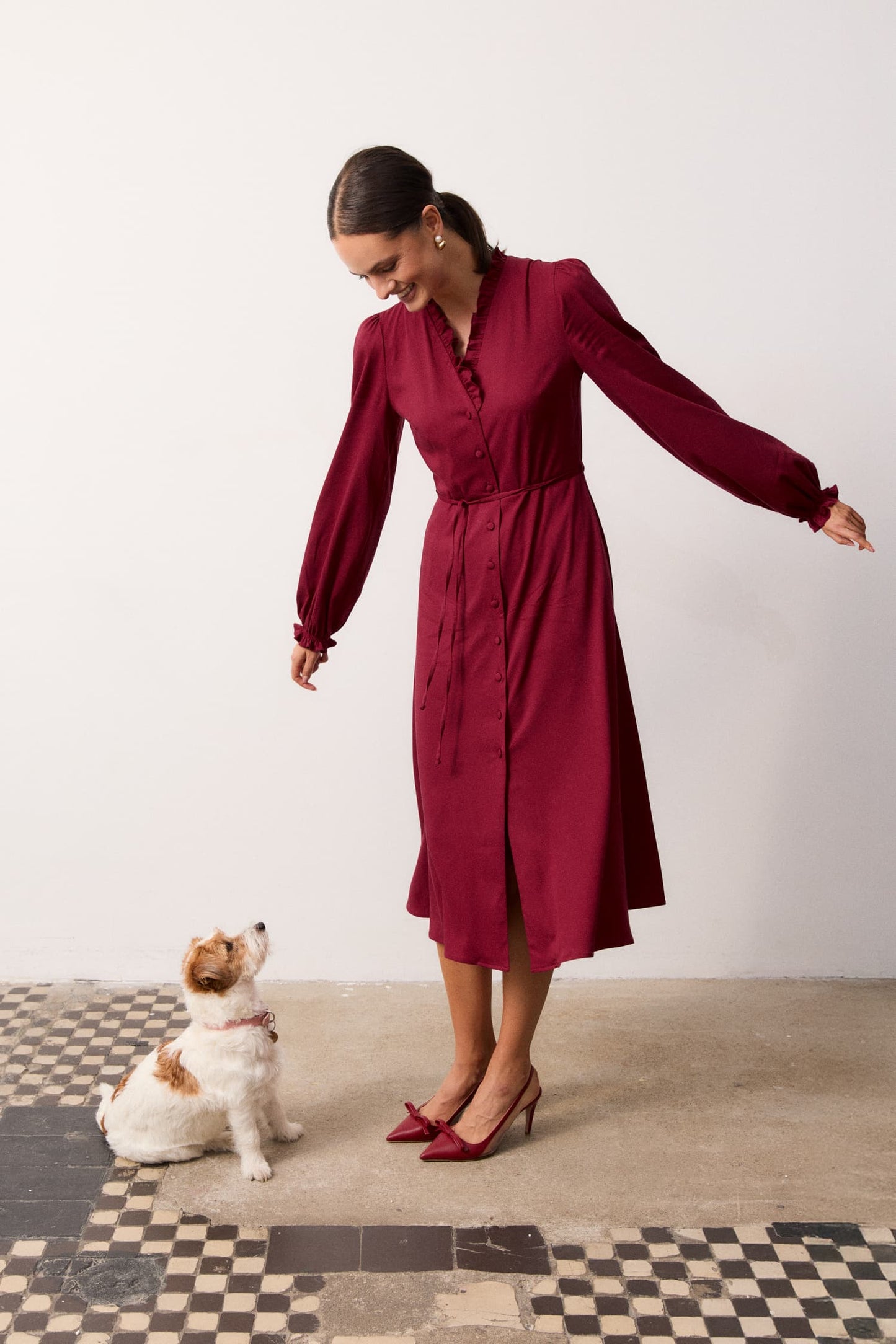 Sukienka midi Bordeaux – bordowa sukienka z długim rękawem i falbanką przy dekolcie, stylizacja świąteczna z czerwonymi szpilkami, modelka z psem – MAG NOLLY