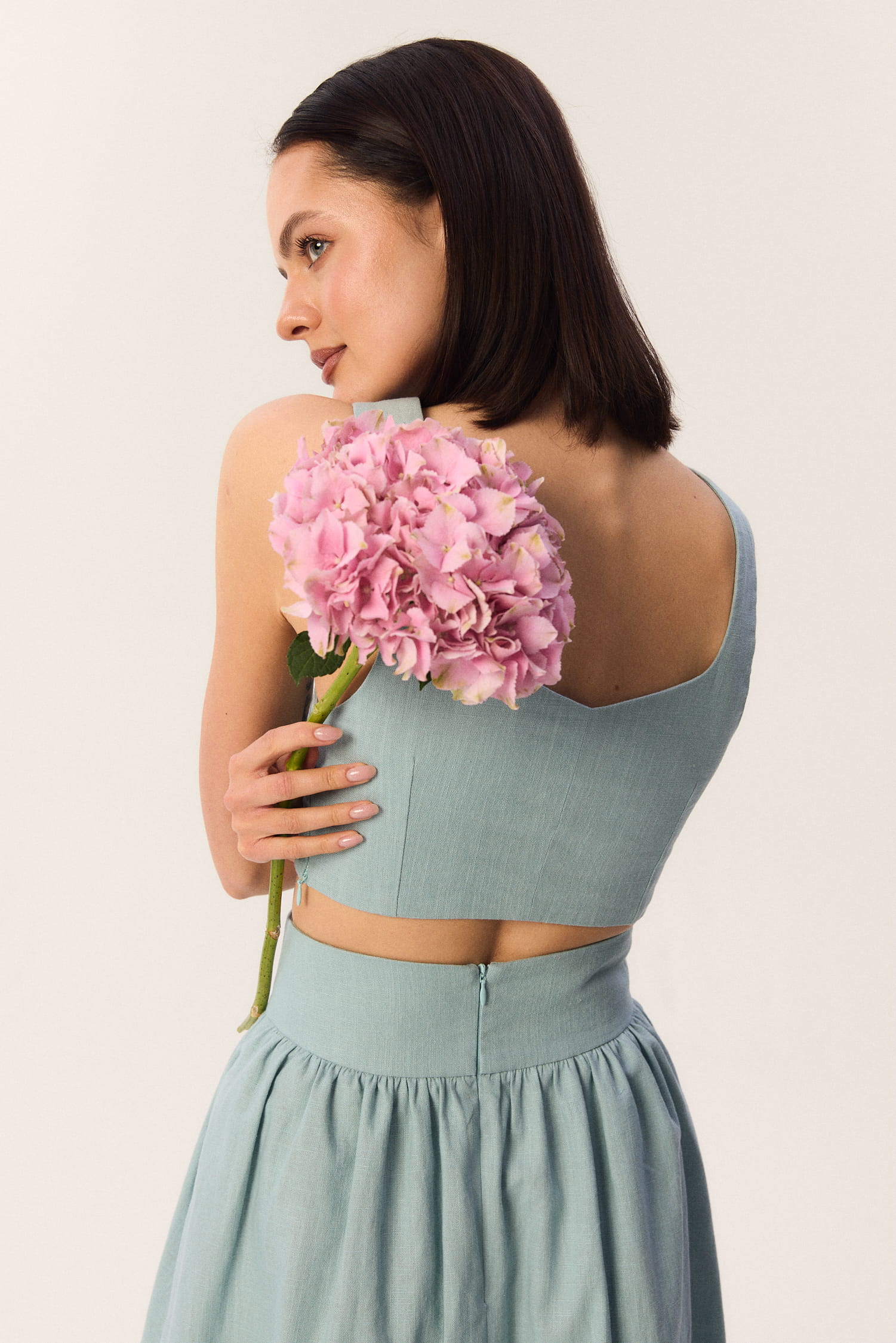 Modelka w topie Milo w kolorze turkusowym, widok z tyłu z różową hortensją, letnia stylizacja ze spódnicą Sandrelli.