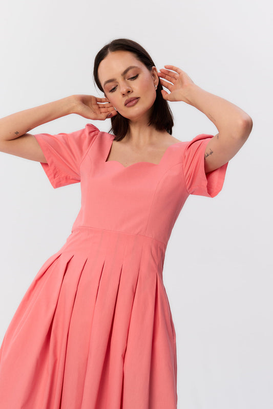 Zbliżenie na rzeźbiarski dekolt w kształcie fali w koralowej sukience midi MAG NOLLY, z krótkim rękawem i zakładkami w talii.