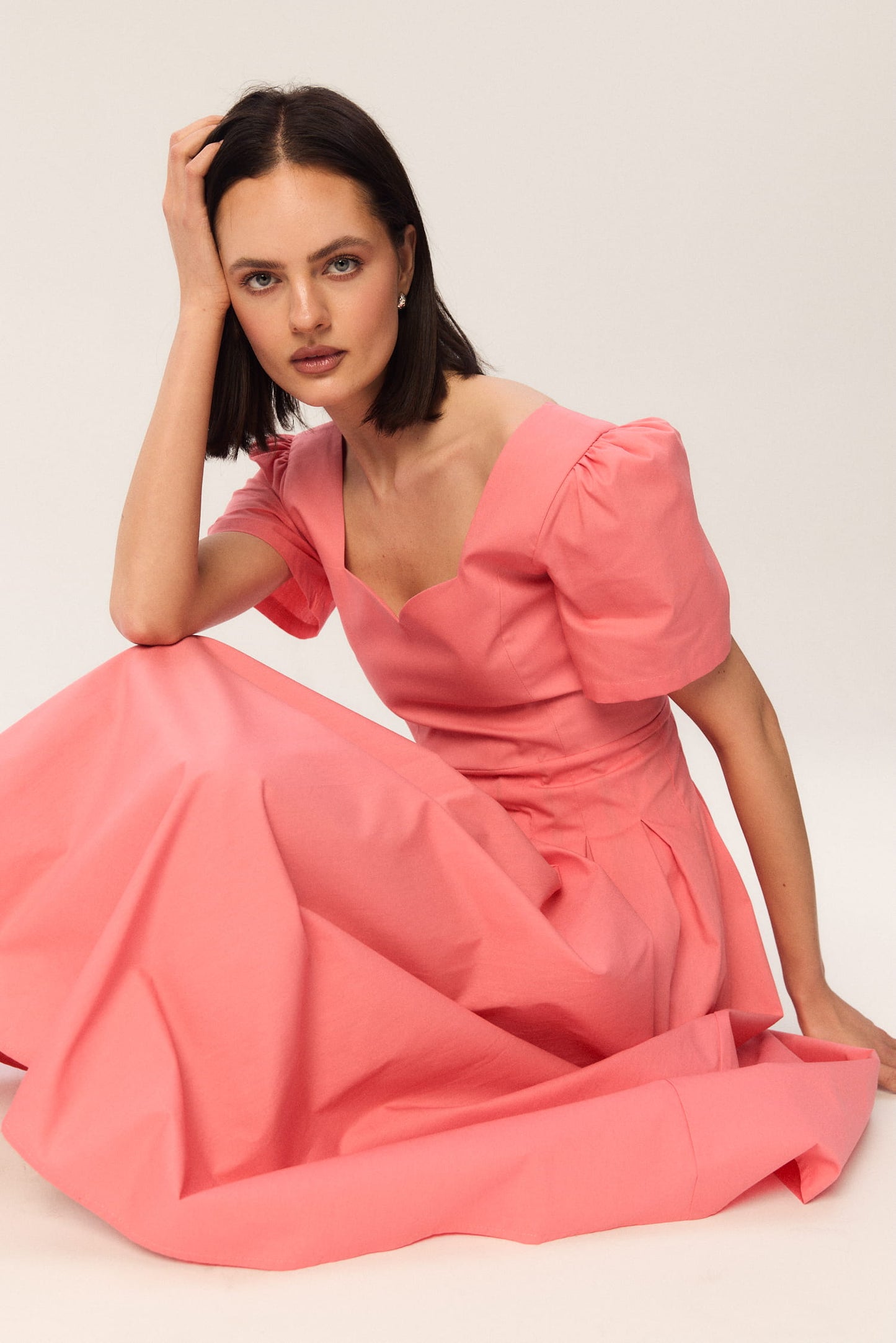 Modelka w koralowej sukience midi MAG NOLLY z falującym dekoltem, siedząca w artystycznej pozie, detal tkaniny i kobiecej sylwetki.
