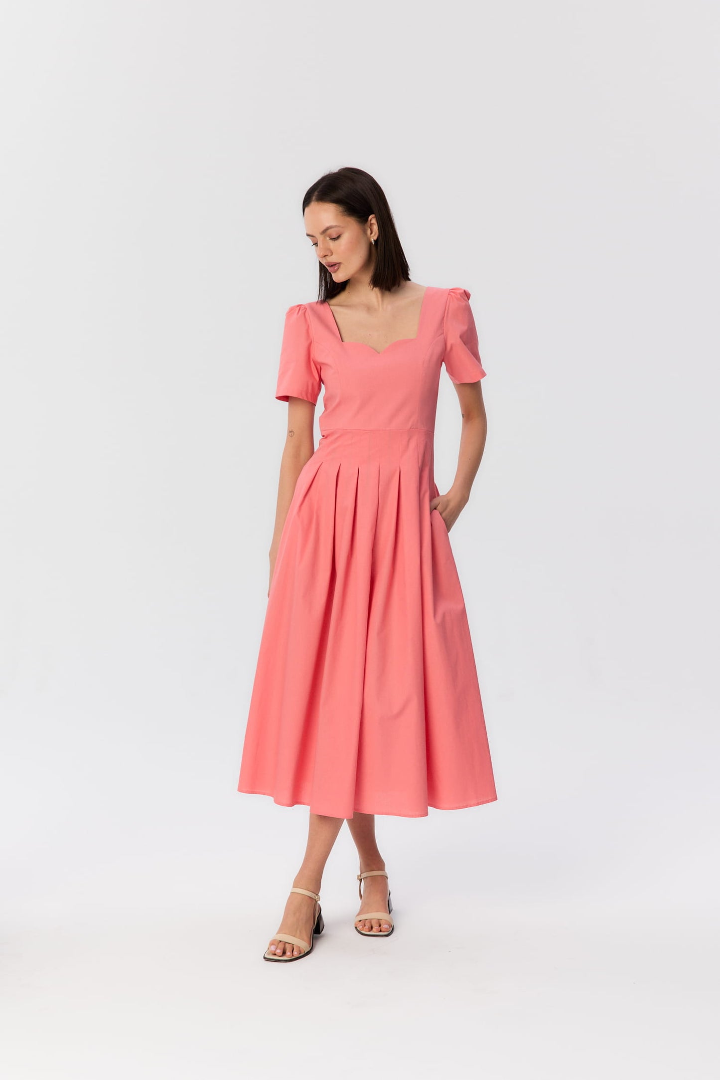 Modelka w koralowej sukience midi MAG NOLLY z falowanym dekoltem i zakładkami, kobieca sylwetka w lekkim ruchu.
