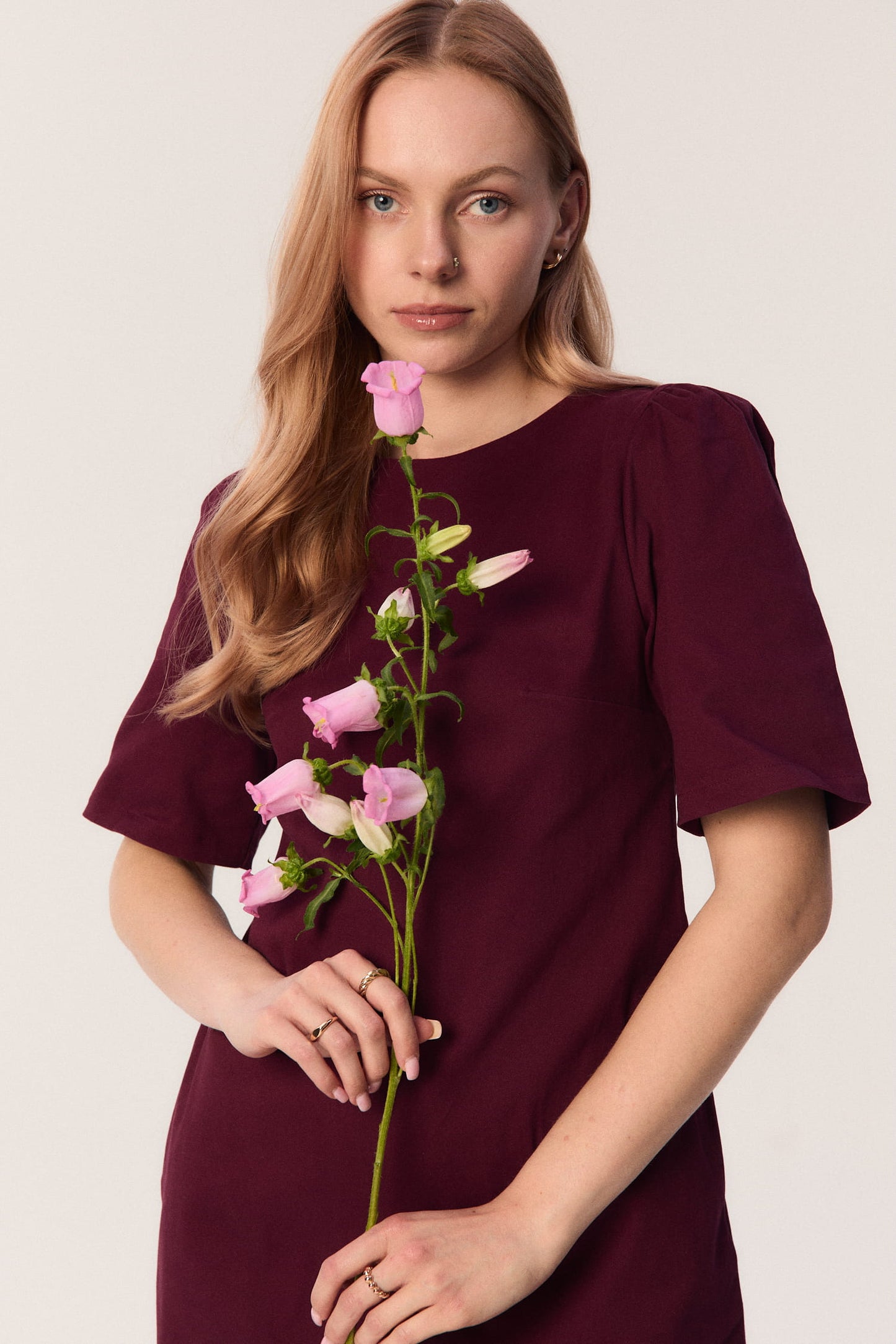 Modelka w bordowej sukience Riva z krótkim rękawem i kwiatem w dłoni – sukienka mini MAG NOLLY.