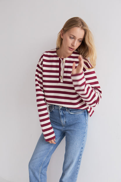 Bluzka Arla Mag Nolly – Bordeaux Milk, paski bordeaux i ecru, guziki przy dekolcie, modelka z ręką w ruchu, niebieska pleciona torebka, stylizacja z jeansami
